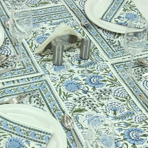 Puede incluir: Un mantel blanco y servilletas a juego con un patrón floral repetitivo en azul y verde. El servicio de mesa incluye platos de cerámica blanca, cubiertos de acero inoxidable, copas de cristal transparente y un servilletero con salero y pimentero.
