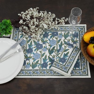 Op de afbeelding: Een rechthoekige placemat met een witte achtergrond en een herhalend blauw en groen bloemmotief, omrand door een bijpassende bloemenrand. Een gevouwen bijpassend servet ligt op de placemat, vergezeld door een wit bord, bestek en een glas.