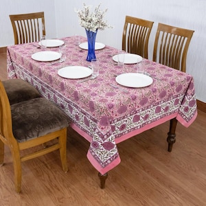 Beautiful hand block printed cotton tablecloth featuring floral patterns with pink and red accents. This vibrant Indian dining table cover adds a touch of bohemian charm to your kitchen or dining space. Ideal for everyday use, festive meals, or special occasions like housewarmings, weddings, baby showers, or dinner parties. Styled on a wooden table with white plates, cutlery, glasses, and a centerpiece flower vase.