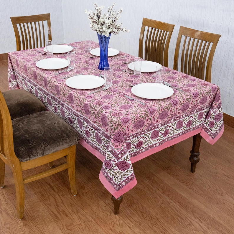 Beautiful hand block printed cotton tablecloth featuring floral patterns with pink and red accents. This vibrant Indian dining table cover adds a touch of bohemian charm to your kitchen or dining space. Ideal for everyday use, festive meals, or special occasions like housewarmings, weddings, baby showers, or dinner parties. Styled on a wooden table with white plates, cutlery, glasses, and a centerpiece flower vase.