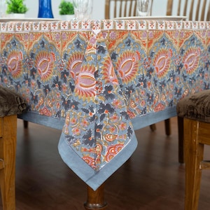 Könnte beinhalten: Rechteckige Tischdecke mit floralem Muster in Blau-, Orange- und Rosatönen. Die Tischdecke ist über einem Holztisch drapiert. Das Design zeigt große, stilisierte Blumen und Blätter. Die Kanten des Stoffes sind sichtbar.
