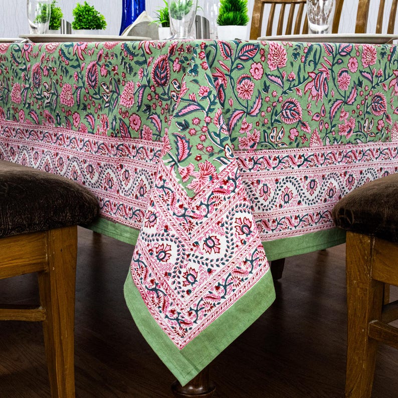 Beautiful hand block printed cotton tablecloth featuring floral patterns with pink and green accents. This vibrant Indian dining table cover adds a touch of bohemian charm to your kitchen or dining space. Ideal for everyday use, festive meals, or special occasions like housewarmings, weddings, baby showers, or dinner parties. Styled on a wooden table with white plates, cutlery, glasses, and a centerpiece flower vase.