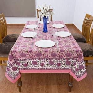 Beautiful hand block printed cotton tablecloth featuring floral patterns with pink and red accents. This vibrant Indian dining table cover adds a touch of bohemian charm to your kitchen or dining space. Ideal for everyday use, festive meals, or special occasions like housewarmings, weddings, baby showers, or dinner parties. Styled on a wooden table with white plates, cutlery, glasses, and a centerpiece flower vase.