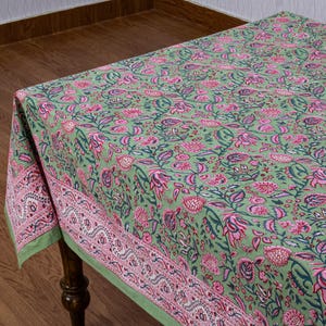 Beautiful hand block printed cotton tablecloth featuring floral patterns with pink and green accents. This vibrant Indian dining table cover adds a touch of bohemian charm to your kitchen or dining space. Ideal for everyday use, festive meals, or special occasions like housewarmings, weddings, baby showers, or dinner parties. Styled on a wooden table with white plates, cutlery, glasses, and a centerpiece flower vase.