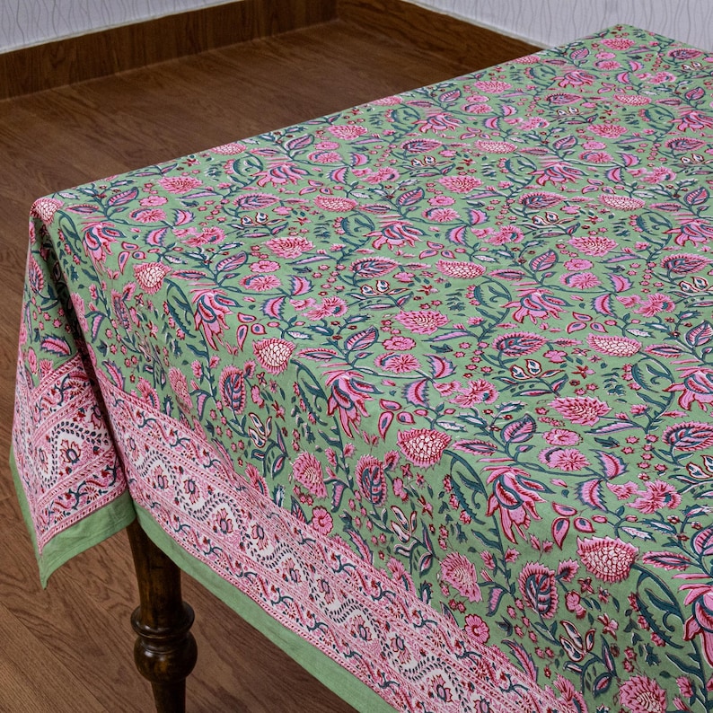 Beautiful hand block printed cotton tablecloth featuring floral patterns with pink and green accents. This vibrant Indian dining table cover adds a touch of bohemian charm to your kitchen or dining space. Ideal for everyday use, festive meals, or special occasions like housewarmings, weddings, baby showers, or dinner parties. Styled on a wooden table with white plates, cutlery, glasses, and a centerpiece flower vase.