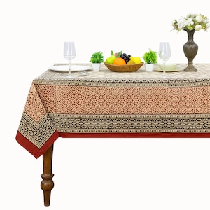 May include: A rectangular tablecloth featuring a repeating geometric pattern in shades of rust, cream, and black. The fabric has a wide border with a dark floral motif, finished with a solid rust-colored edge, draped over a wooden table.