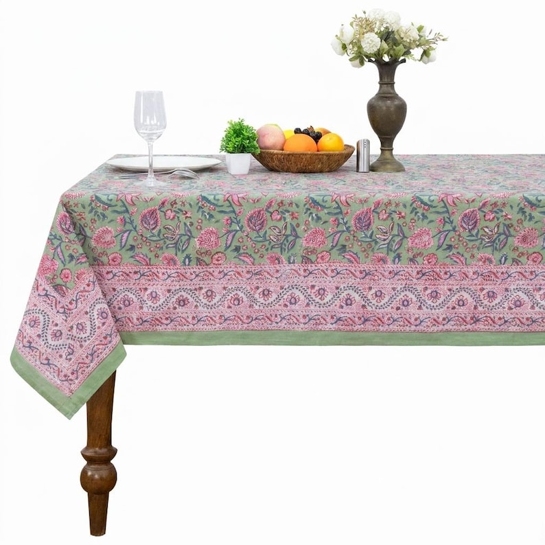 Beautiful hand block printed cotton tablecloth featuring floral patterns with pink and green accents. This vibrant Indian dining table cover adds a touch of bohemian charm to your kitchen or dining space. Ideal for everyday use, festive meals, or special occasions like housewarmings, weddings, baby showers, or dinner parties. Styled on a wooden table with white plates, cutlery, glasses, and a centerpiece flower vase.