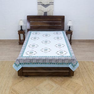 May include: A white bedspread with a floral pattern in teal and gray, framed by a teal and gray border. The bedspread is on a dark wooden bed frame, with matching nightstands and lamps on either side.