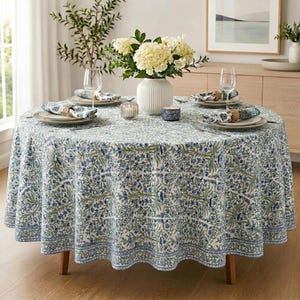 Può includere: Un tavolo da pranzo rotondo coperto da una tovaglia bianca con un motivo floreale blu e verde. Il tavolo è apparecchiato con quattro piatti bianchi, ciascuno con un tovagliolo arrotolato, quattro calici trasparenti e un vaso bianco centrale.
