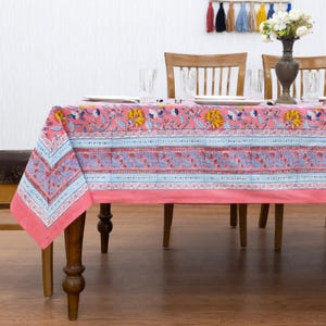 Pode incluir: Uma toalha de mesa rosa floral com um padrão repetido de flores amarelas, folhas azuis e detalhes em vermelho. A toalha de mesa é retangular e cobre uma mesa de madeira. A mesa está posta com pratos, copos e um vaso de flores brancas.