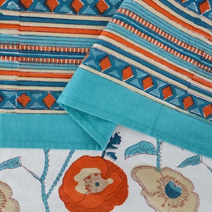 May include: A close-up of folded turquoise fabric with a geometric pattern of orange, blue, and beige stripes and diamond shapes. The fabric is placed over a white floral print with orange and beige flowers, and blue stems and leaves. The theme is home decor.