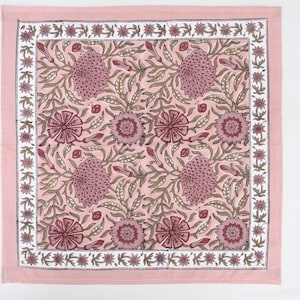 Puede incluir: Un camino de mesa de algodón rosa con estampado floral. El estampado presenta flores y hojas rosas y marrones sobre un fondo rosa claro. El camino de mesa tiene un borde blanco con un patrón de estrellas repetido.