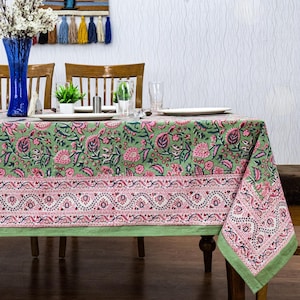 Beautiful hand block printed cotton tablecloth featuring floral patterns with pink and green accents. This vibrant Indian dining table cover adds a touch of bohemian charm to your kitchen or dining space. Ideal for everyday use, festive meals, or special occasions like housewarmings, weddings, baby showers, or dinner parties. Styled on a wooden table with white plates, cutlery, glasses, and a centerpiece flower vase.