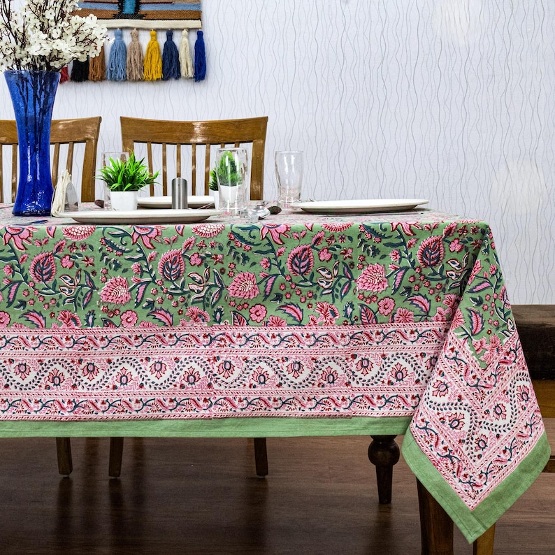 Beautiful hand block printed cotton tablecloth featuring floral patterns with pink and green accents. This vibrant Indian dining table cover adds a touch of bohemian charm to your kitchen or dining space. Ideal for everyday use, festive meals, or special occasions like housewarmings, weddings, baby showers, or dinner parties. Styled on a wooden table with white plates, cutlery, glasses, and a centerpiece flower vase.