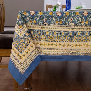 May include: A rectangular tablecloth featuring a blue background with a repeating floral pattern in yellow, cream, and green. The design includes horizontal bands of intricate botanical motifs and a solid blue border along the edges of the fabric.