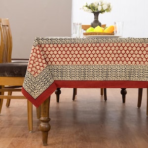 May include: A rectangular tablecloth featuring a repeating cream and dark brown leaf pattern on a deep red background, with a wide dark brown border containing a geometric motif and a solid red hem, draped over a wooden dining table.