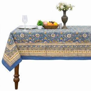 May include: A rectangular tablecloth featuring a blue base with a repeating floral pattern in yellow, green, and white. The fabric has a wide decorative border with intricate botanical motifs in muted tones, finished with a solid blue hem.