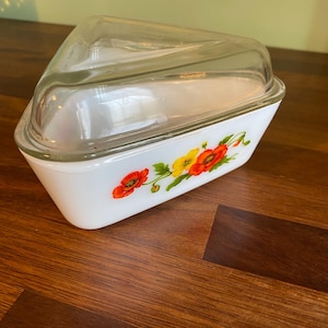 May include: A white, triangular casserole dish with a clear glass lid. The dish has a floral design with red and yellow poppies. The dish is on a wooden surface.