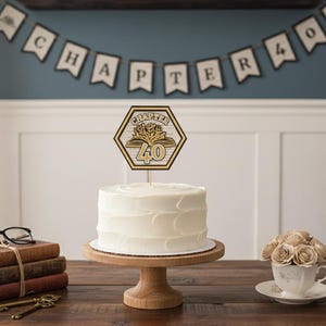 May include: A white frosted cake on a wooden cake stand, topped with a Chapter 40 cake topper. The scene includes a banner that reads "CHAPTER 40", books, keys, glasses, and a teacup with flowers. The cake is the focal point.