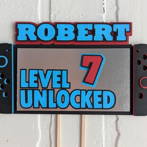 May include: A black and silver cake topper in the shape of a game console. The topper reads "ROBERT" in blue letters with a red outline, and "LEVEL UNLOCKED 7" in blue with a red outline.