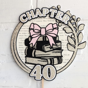 May include: A gold glitter cake topper with a stack of books, a pink bow, and the words "Chapter 40".