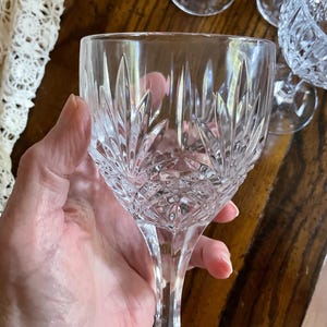 May include: A clear crystal wine glass with a detailed, cut-glass design. The glass features a long stem and a bowl with a pattern of vertical and diamond-shaped cuts. Other crystal glassware is visible in the background.