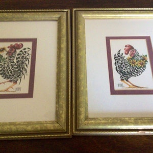 May include: Two framed rooster art prints. Each print features a rooster with black and white patterned feathers, red comb, and yellow flowers. The prints are set in gold frames with a white mat and a purple border.