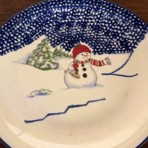 May include: White ceramic plate with a winter scene. A snowman wearing a red hat and scarf stands in the snow, with a green Christmas tree in the background. The rim of the plate has a blue dotted pattern.