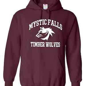 May include: A maroon hooded sweatshirt with the text "MYSTIC FALLS" arched above a white wolf head graphic, and "TIMBER WOLVES" below. The sweatshirt has a front pocket and a drawstring hood.