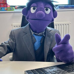 May include: A purple puppet wearing a gray suit, a checkered shirt, and a blue tie, seated at a desk. The puppet has black hair and is gesturing with its right hand. A keyboard is in front of the puppet.