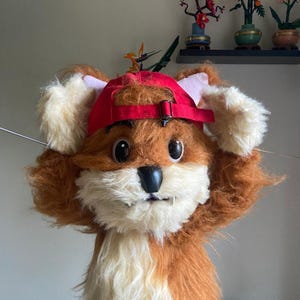 May include: A plush fox character wearing a red baseball cap. The fox has brown and cream-coloured fur, large black eyes, and a black nose. The fox is holding its ears with its hands.