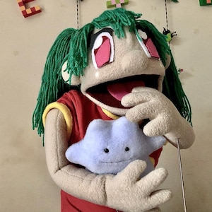 Könnte beinhalten: Eine handgefertigte Marionette mit grünen Garnhaaren, großen Augen mit rosa Rändern und einem roten Hemd mit gelbem Besatz. Die Marionette hält ein hellblaues sternförmiges Plüschtier. Der Mund der Marionette ist geöffnet.