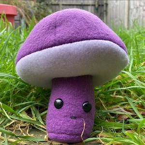 May include: A plush mushroom toy with a purple cap and stem, a light gray underside, and a smiling face with black button eyes. The toy is set against a backdrop of green grass.