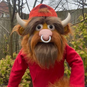 Puede incluir: Una marioneta artesanal con apariencia de toro. Presenta una gorra roja, una camisa roja y un pelaje largo y desgreñado, marrón y naranja. La marioneta tiene grandes cuernos blancos, un aro nasal rosa y una larga barba marrón.