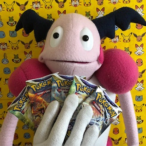 Könnte beinhalten: Eine rosa Puppe mit schwarzen Flügeln und roten Armen hält mehrere Pokémon Trading Card Game Booster Packs. Der Hintergrund ist gelb mit einem sich wiederholenden Muster verschiedener Pokémon-Charaktere.