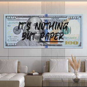 Op de afbeelding: Een grote rechthoekige canvas print van een Amerikaanse $100 biljet met de tekst "IT'S NOTHING BUT PAPER" over het beeld. Het kunstwerk toont een portret van Benjamin Franklin en wordt gepresenteerd in een moderne woonkamer.