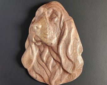 May include: A light brown wooden wall carving featuring a detailed relief of a dog head with long, drooping ears and a wrinkled face. The natural wood grain is visible throughout the surface, highlighting the sculpted contours of the canine features.