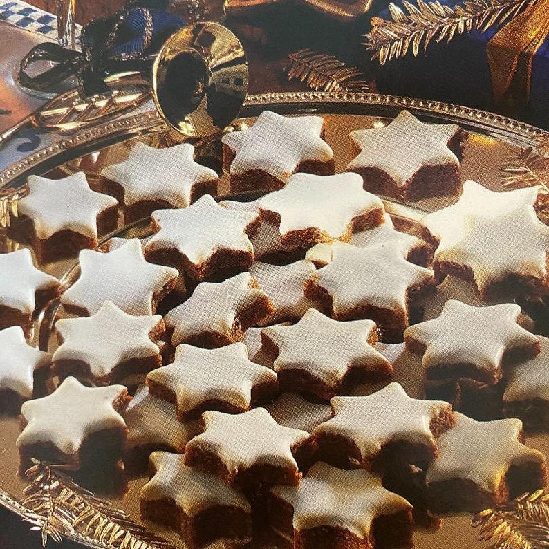 K&ouml;nnte beinhalten: Ein silbernes Tablett mit sternf&ouml;rmigen Keksen. Jeder Keks hat einen hellbraunen Boden und ist mit wei&szlig;em Zuckerguss bedeckt. Die Kekse sind leicht &uuml;berlappend angeordnet und bilden eine ansprechende Pr&auml;sentation.