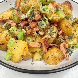 Puede incluir: Un plato de patatas asadas de color marrón dorado, adornadas con eneldo verde fresco y cebolletas en rodajas, y trozos de bacon cocido. Las patatas son el foco principal, con las hierbas y el bacon añadiendo color y textura. El plato tiene un borde negro.