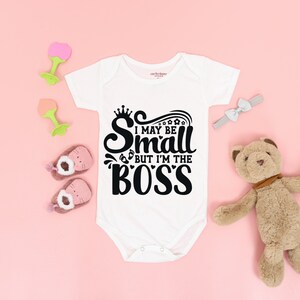Könnte beinhalten: Weißer Baby-Strampler mit schwarzem Text, der "I may be small but I'm the BOSS" lautet. Der Text ist in einer stilisierten Schriftart mit einer Krone über dem ersten Buchstaben.