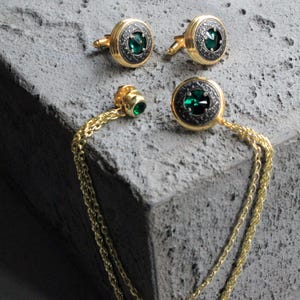 May include: A set of gold-toned cufflinks and a tie pin with a matching design. Each piece features a dark green gemstone set within a circular, ornate frame. The tie pin is connected to a gold chain.