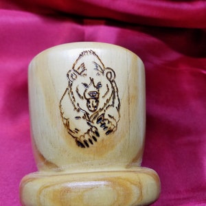 May include: A light-colored wooden cup with a detailed, burnt-wood image of a bear. The bear is centered on the cup, with its mouth open and claws visible. The cup has a rounded base and a smooth finish.