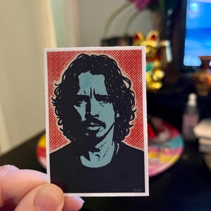 Puede incluir: Una pegatina de estilo pop art con un retrato de un hombre con pelo largo y rizado oscuro y perilla. El retrato está en tonos azules y negros sobre un fondo rojo punteado. La pegatina es rectangular y tiene un borde blanco.