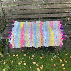 May include: A colourful, rectangular rag rug with a striped pattern. The rug is made from strips of pink, yellow, blue, and white fabric, with fringed edges in pink, purple, and white. It is placed on a wooden bench.