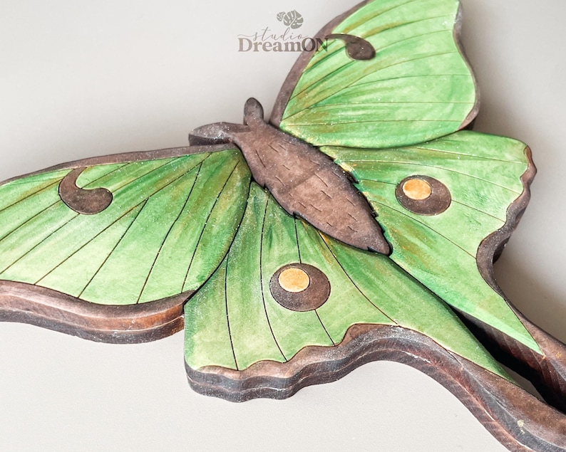 K&ouml;nnte beinhalten: Eine Wandskulptur aus Holz, die einen gr&uuml;nen Luna-Motte mit braunen Akzenten darstellt. Der Falter hat gro&szlig;e Fl&uuml;gel mit augenartigen Markierungen.