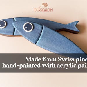 May include: Two blue wooden fish decorations with white and black eye details. The fish are hand-painted with acrylic paint and made from Swiss pine. The text in the image says "Made from Swiss pine & hand-painted with acrylic paint."