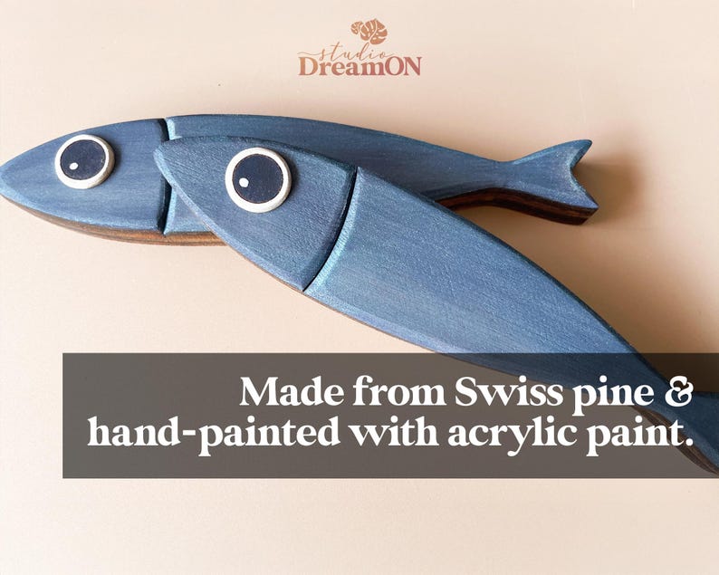 May include: Two blue wooden fish decorations with white and black eye details. The fish are hand-painted with acrylic paint and made from Swiss pine. The text in the image says "Made from Swiss pine & hand-painted with acrylic paint."
