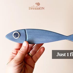 May include: A hand-painted, blue wooden fish with a white-rimmed eye. The fish is a decorative item, with the text "Just 1 fish" in a gray box. The Studio DreamON logo is in the upper left corner.