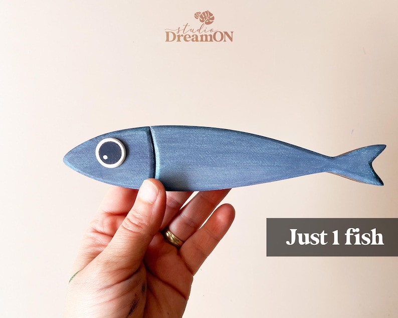 May include: A hand-painted, blue wooden fish with a white-rimmed eye. The fish is a decorative item, with the text "Just 1 fish" in a gray box. The Studio DreamON logo is in the upper left corner.