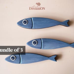 May include: Three wooden fish decorations in varying shades of blue. Each fish has a white and black eye and a detailed tail fin. The text "Bundle of 3" is displayed in a black box. The Studio DreamON logo is in the top right corner.